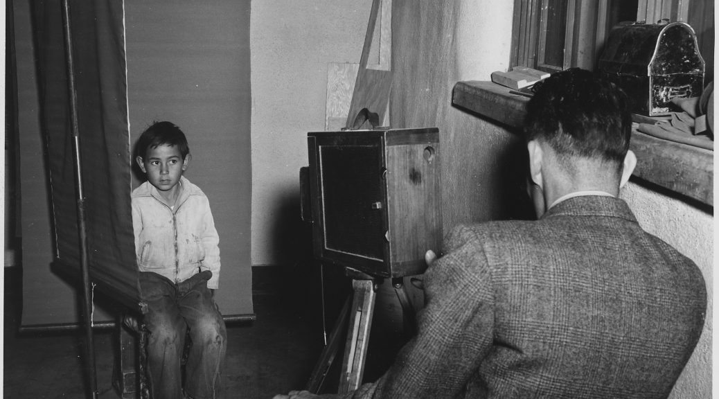 Black and white photograph of boy wearing a light colored jacket and worn out blue jeans, is seated for a photograph in front of a dark photo backdrop. The photographer, a man in a suit is seen from the back, operating a wood box camera on a tripod. To theright is a window with an old fashioned black metal lunchbox resting on the window sill.
