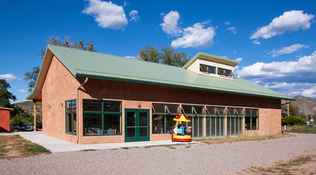 Color photograph of Adobe library building with a plastic children's carousel in bright primary colors in front of the building. In the foreground is a gravel parking lot. Libray building is framed against a bright blue sky with scattered clouds.