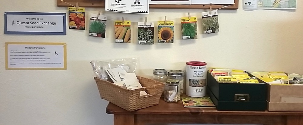 Table with boxes jars and baskets of seed packets and a donation jar. Hanging above these on string with clothesline is additional seed packets. On the wall are instructions for participating in seed exchange.