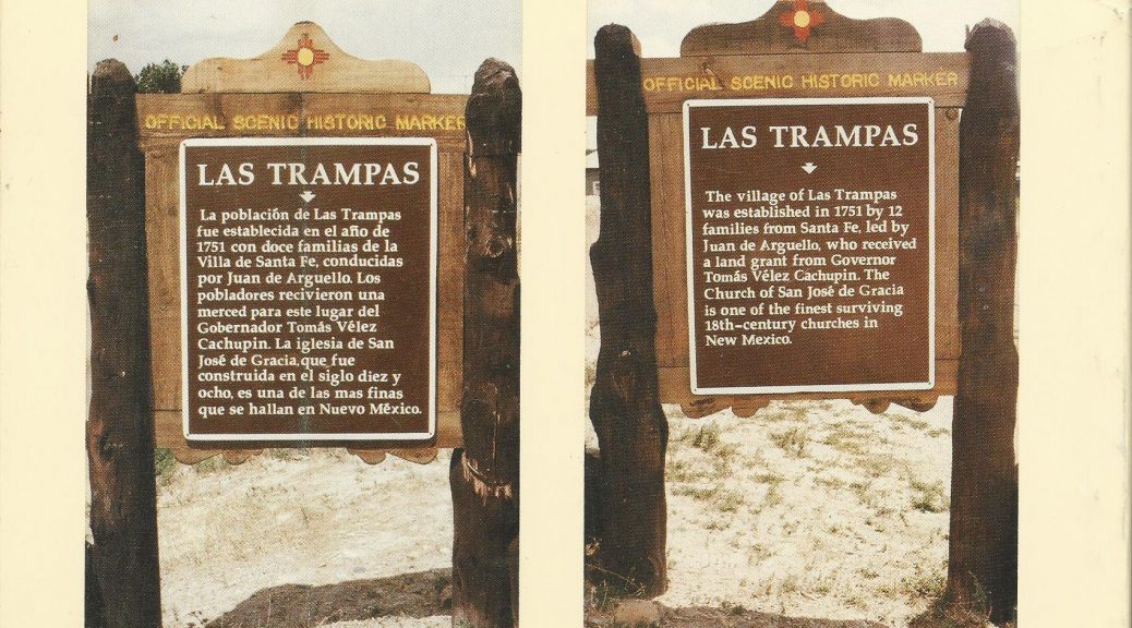 Two photos, side by side, of official scenic markers with historical information printed on metal signage affixed to wood, mounted on rough wooden poles, for the village of Las Trampas. Decorative trim with Zia symbol is attached at the top of both images. The text for the English sign reads: The village of Las Trampas was established in 1751 by 12 families from Santa Fe, led by Juan de Arguello, who received a land grant from governor Tomás Vélez Capuchin. The Church of San José de Gracia is one of the finest surviving 18th century churches in New Mexico. The Spanish sign reads: La poblaciōn de Las Trampas fun establecida en al año de 1751 con doce families de la Villa de Santa Fe, conducidas for Juan de Arguello. Los pobladores recivieron una merced para este lugar del Gobernador Tomás Velez Capuchin. La iglesia de San José de Gracia, que fue construida en el siglo diez y ocho, es una de las las finas que se hallan en Nuevo México.