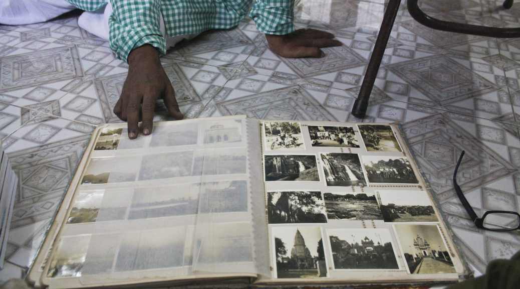 Photo album open to display images, resting on a tile floor near eyeglasses and a chair. A man's hand holds open a page. On the page are black and white photos of scenes of people, buildings and waterfalls.