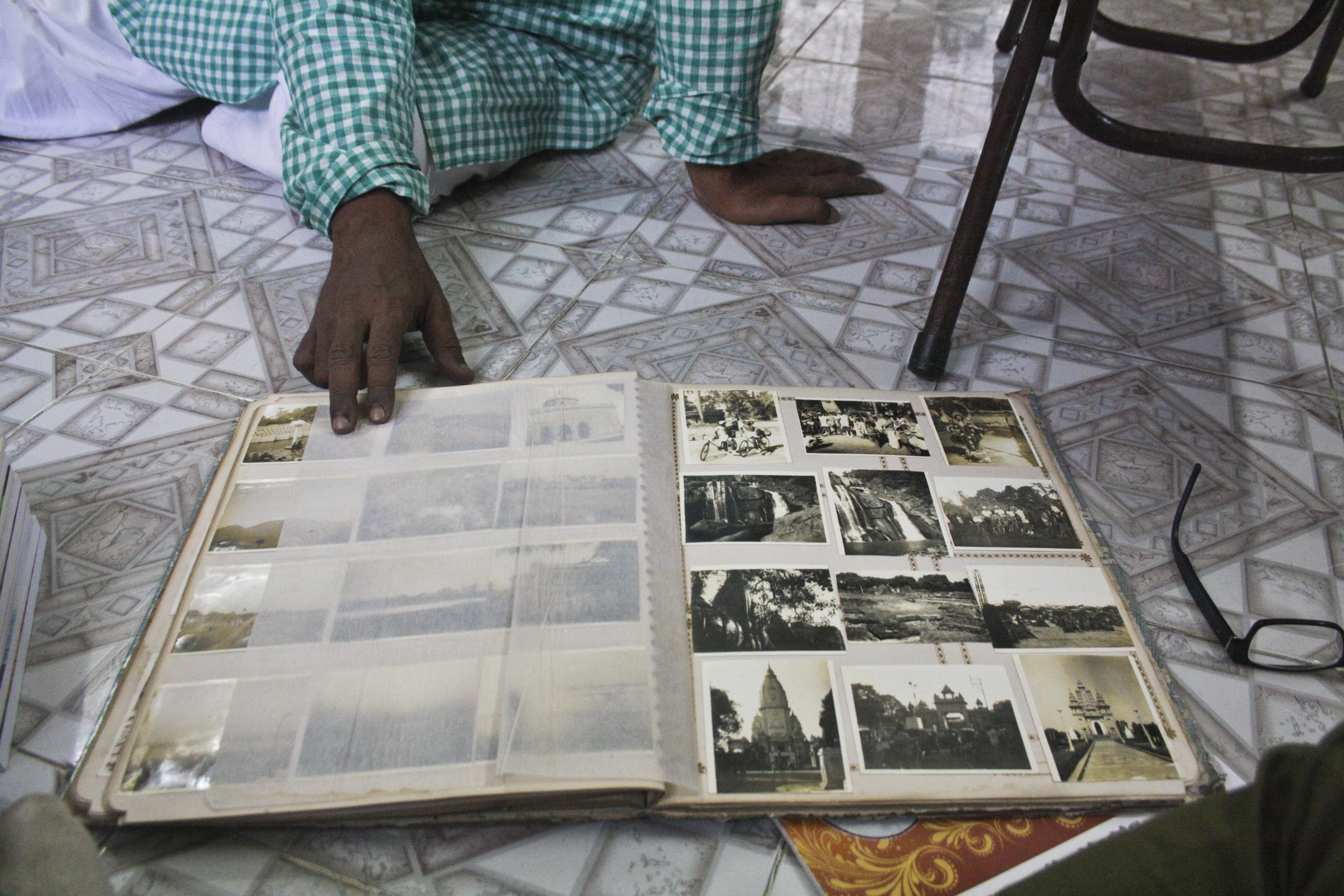 Photo album open to display images, resting on a tile floor near eyeglasses and a chair. A man's hand holds open a page. On the page are black and white photos of scenes of people, buildings and waterfalls.
